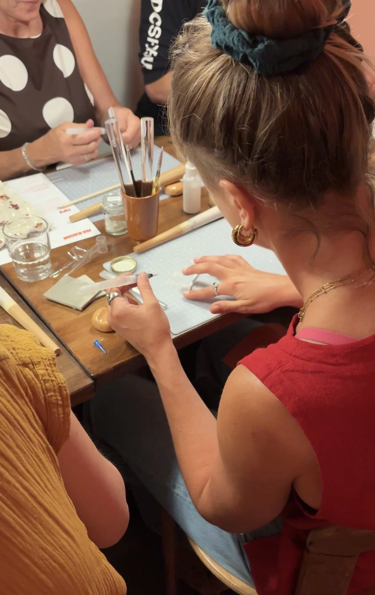Une femme avec des boucles d'oreilles en or, portant une robe sans manches rouge, est assise à une table, utilisant un outil pour appliquer du gel ou du maquillage sur sa main alors que d'autres personnes participent à une activité créative.