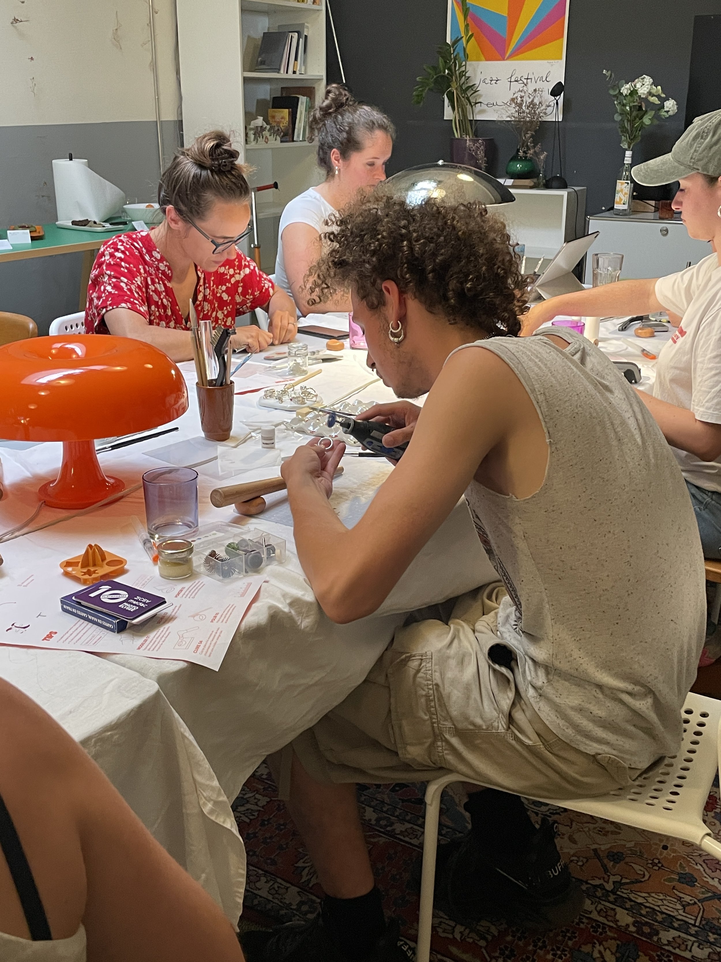 Groupe de personnes assises autour d'une table, travaillant ou faisant du bricolage, dans une pièce décorée avec un poster coloré et des plantes.