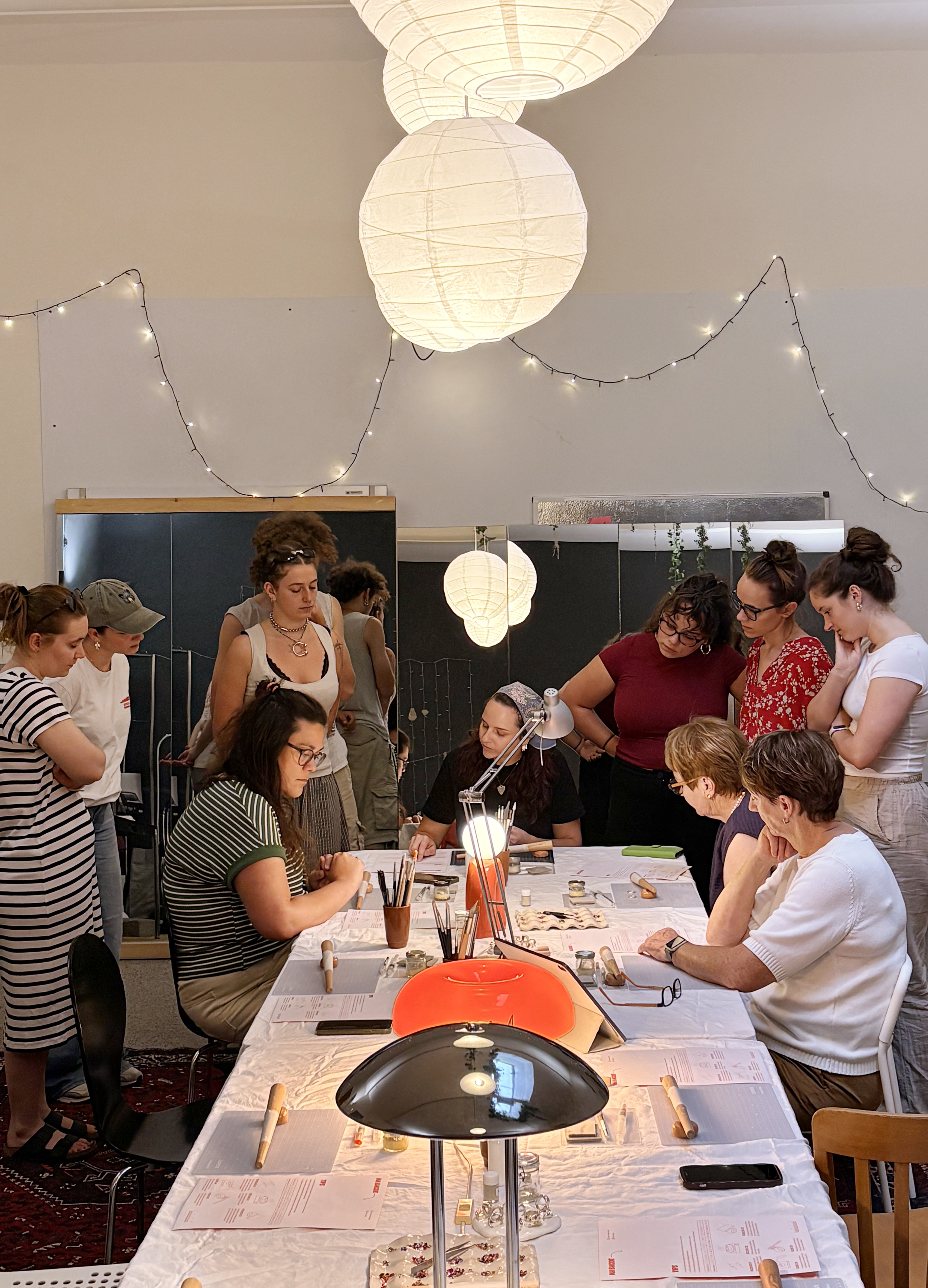 Groupes de personnes rassemblées autour d'une table lors d'un atelier créatif ou artisanal dans une salle décorée avec des lampions en papier et des guirlandes lumineuses.