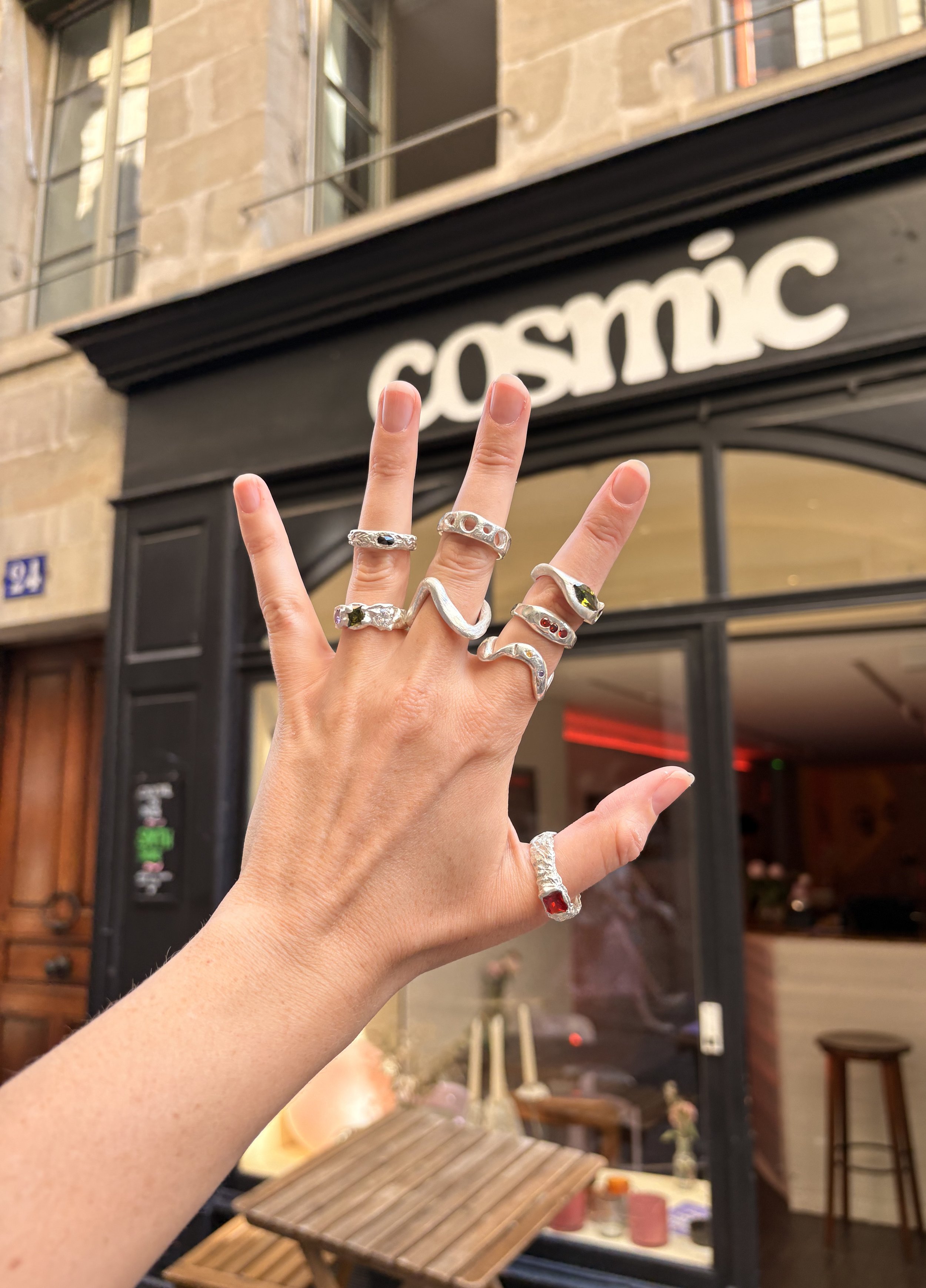 Une main portant plusieurs bagues en argent, devant un bâtiment avec une enseigne 'COSMIC'.
