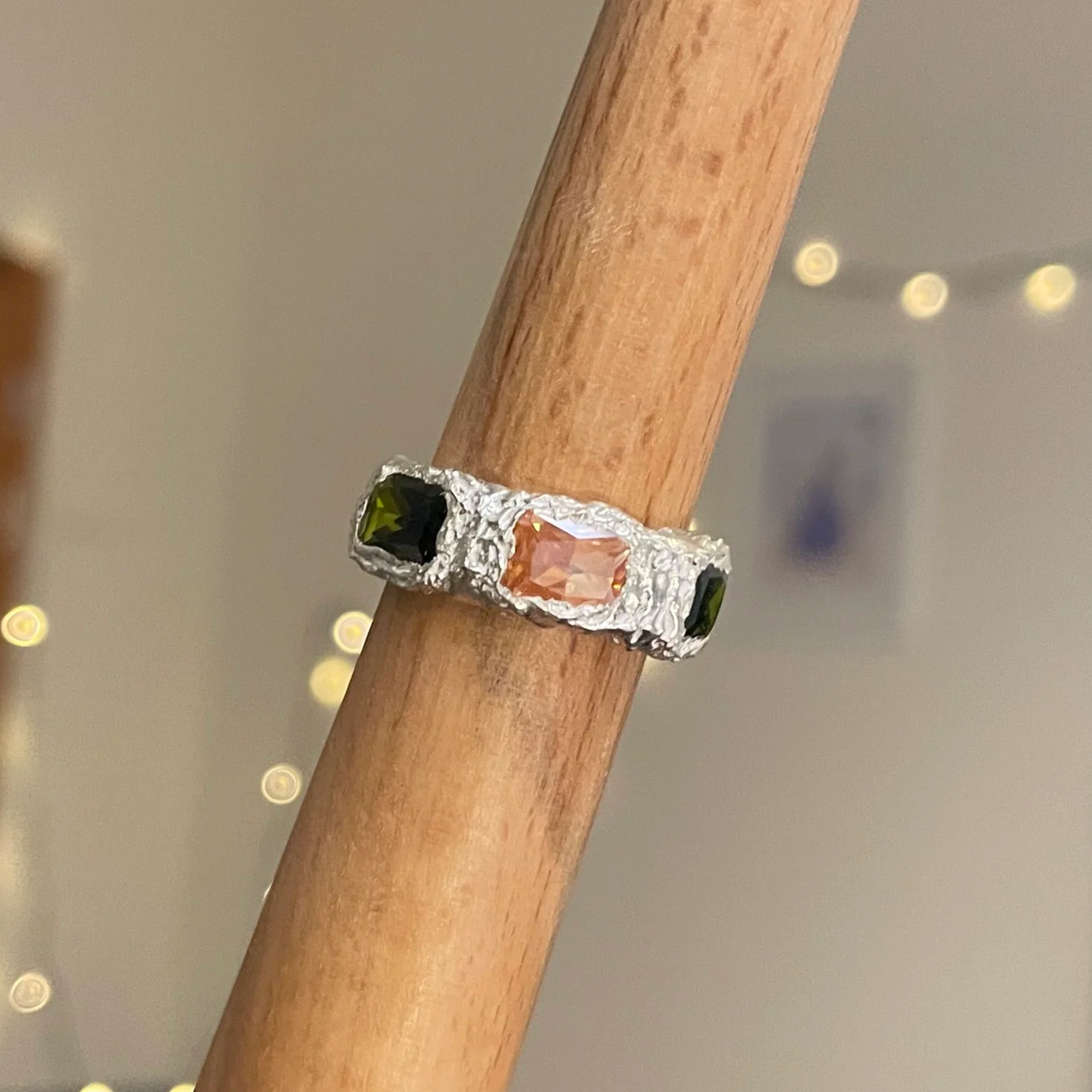 Une bague avec trois pierres précieuses, une centrale orange et deux vertes, sur un morceau de bois, avec un fond flou de lumières chaudes.