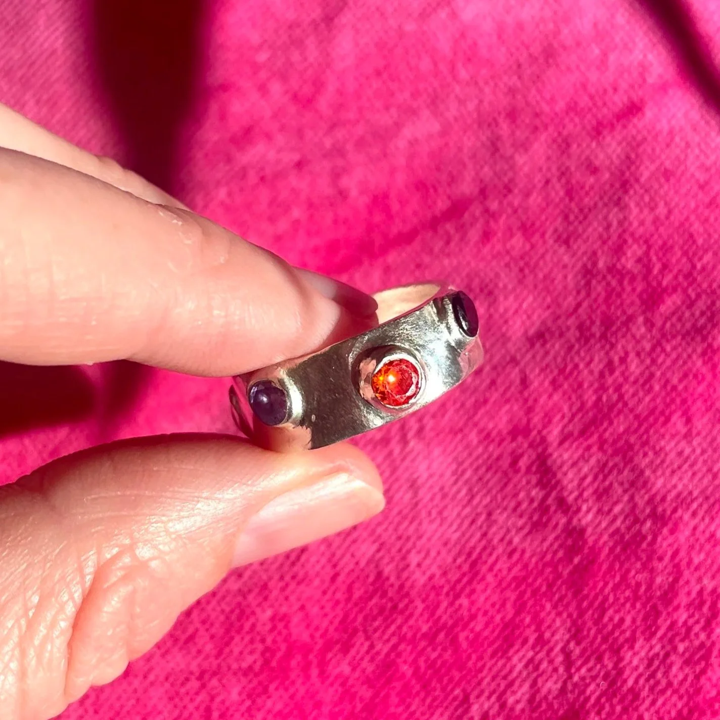 Une personne tenant une bague en métal avec une pierre rouge au centre et deux pierres bleues de chaque côté, contre un fond rose.
