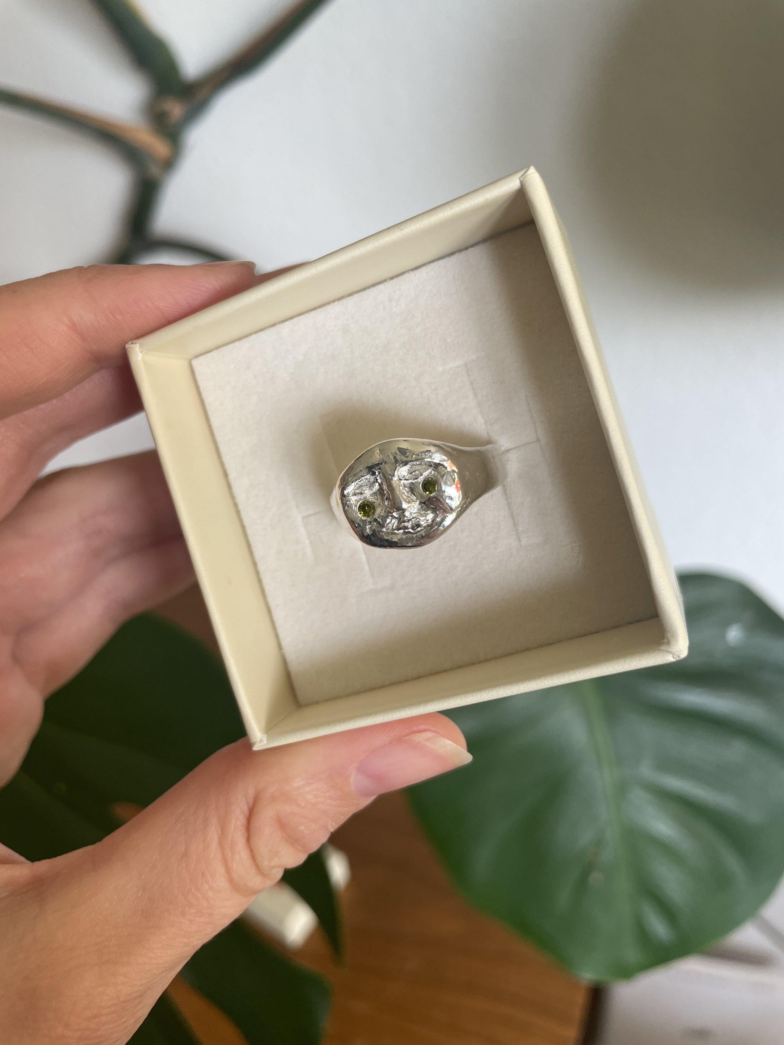 Une bague en argent avec deux pierres vertes est présentée dans une petite boîte crème.
