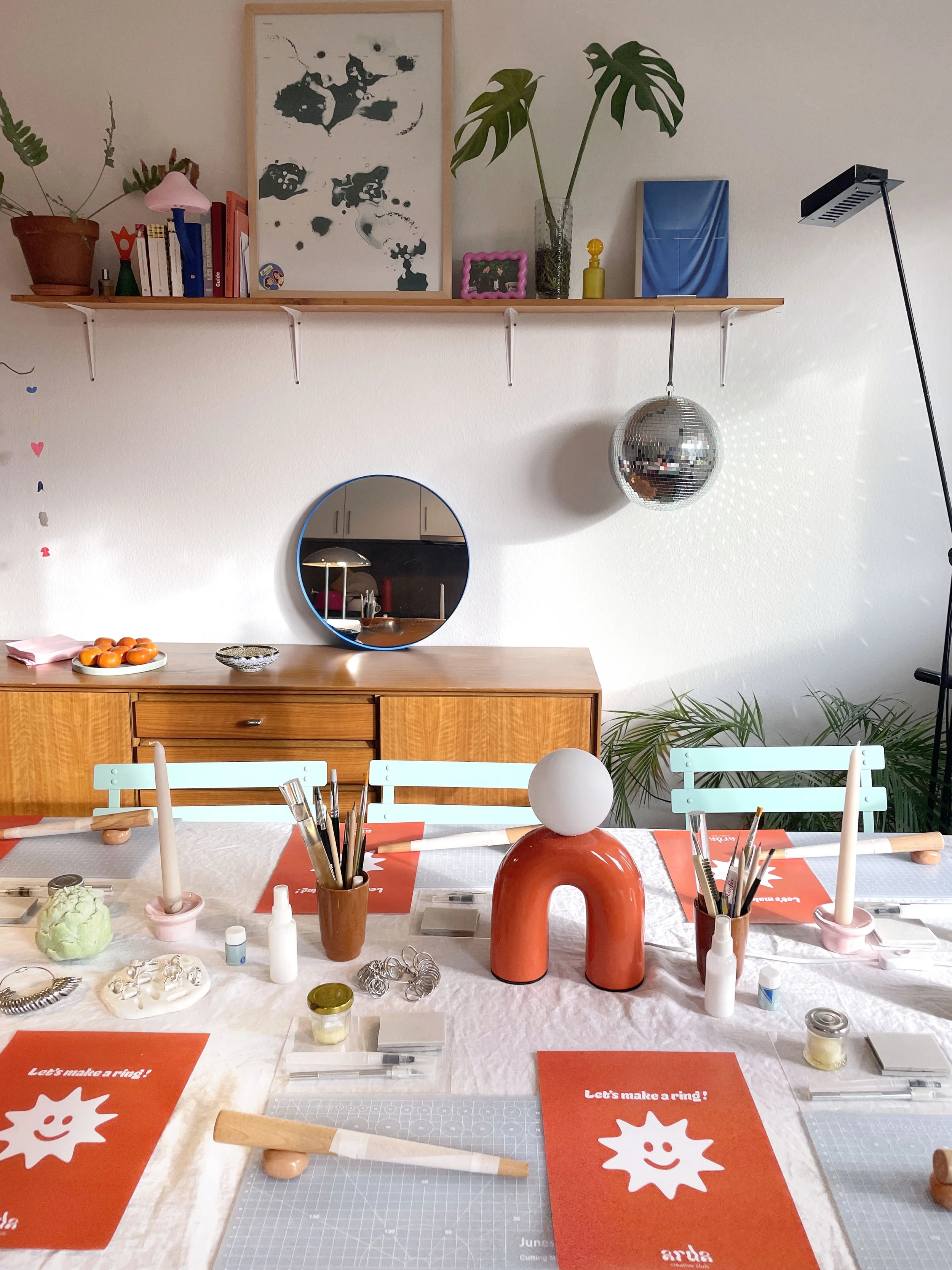 Une table de bricolage pour faire des bagues avec des matériaux, outils, et fiches d'instructions. Des fiches rouges avec l'inscription 'Let's make a ring !' sont disposées sur la table.