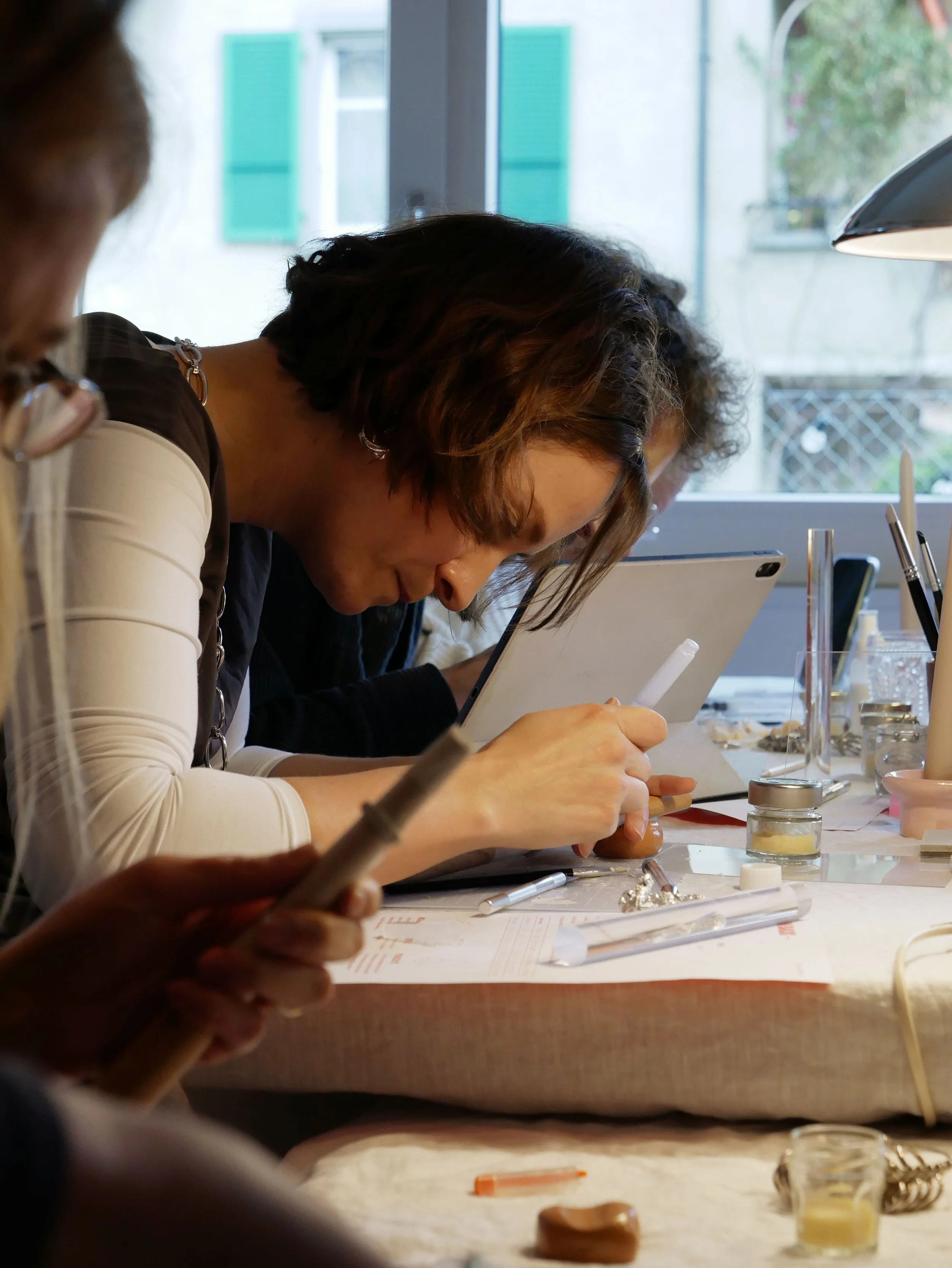 Des femmes travaillent dans un atelier de bijoux, concentrées sur leurs créations avec divers outils et matériaux sur la table.