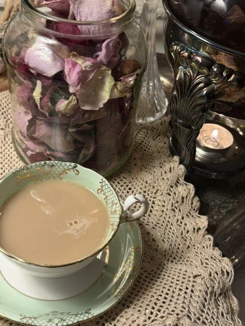 A cup of tea or coffee with cream on a saucer, a glass jar filled with dried rose petals, and a decorative oil or candle holder with a lit candle on a crocheted cloth.