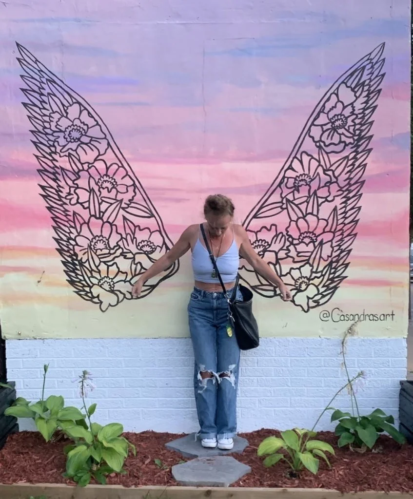 A woman standing in front of a colorful mural with butterfly wing designs. She is wearing a white crop top and ripped jeans, with her arms outstretched and looking down. There are green plants in pots at her feet.