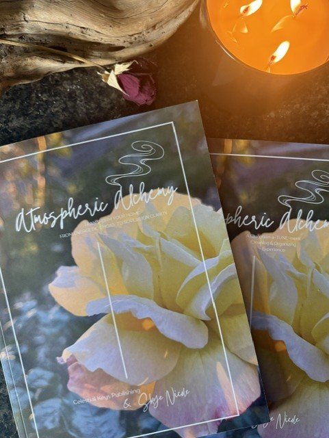 Two pamphlets titled 'Atmospheric Alchemy' with a yellow rose on the cover, a lit candle in an orange holder, a small piece of driftwood, and a dried flower on a dark surface.