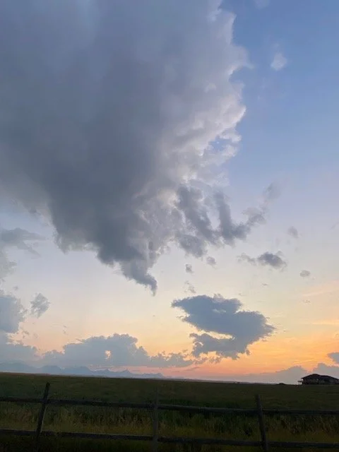 Sky with large dark clouds and a hint of sunset near the horizon, over a field with a fence.