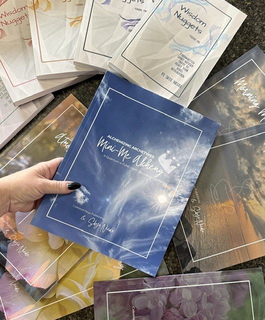 Hand holding a blue book titled "Alchemical Archetypes Mini-Moon Readings" surrounded by various other colorful books on a dark surface.