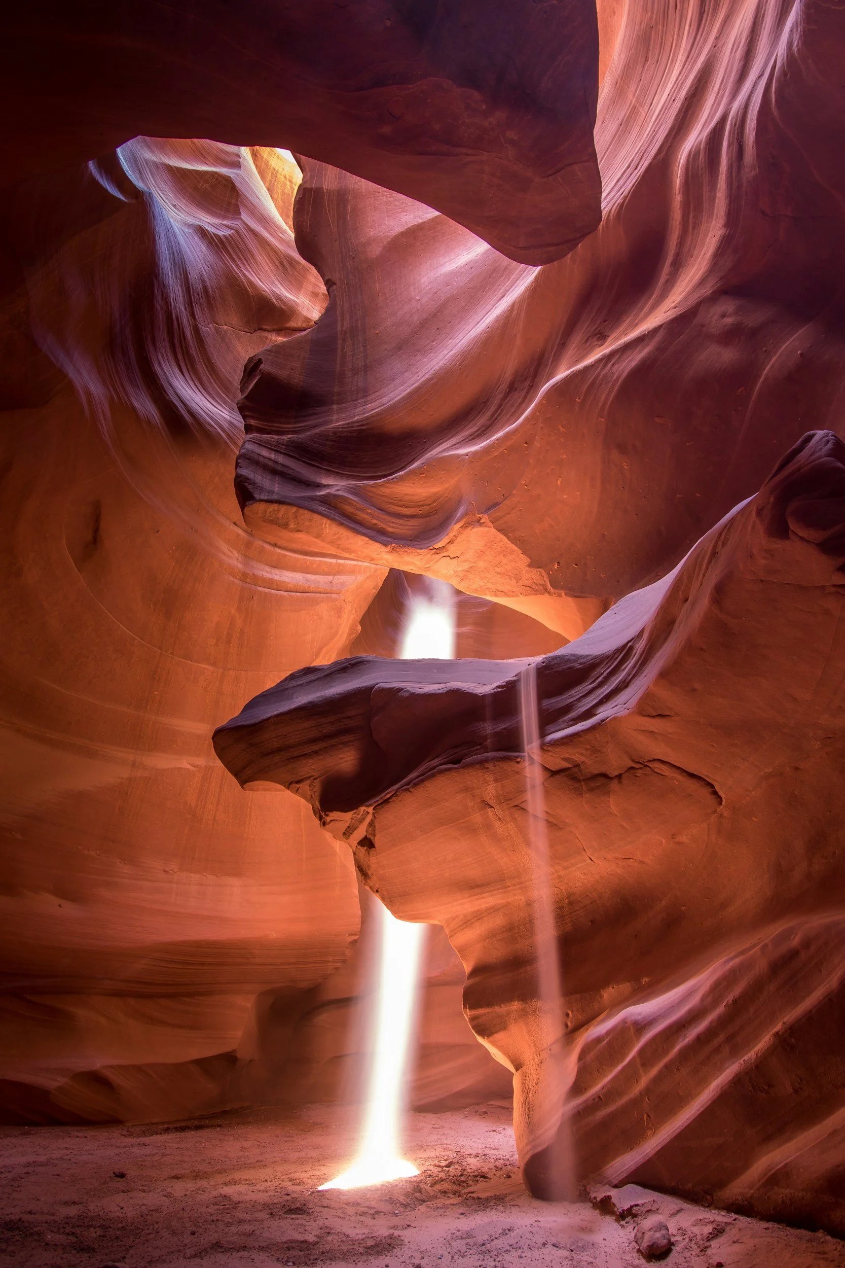 Light beams entering the Grand Canyon, illuminating the smooth, layered red and orange sandstone walls, with a sandy floor and a small rock.