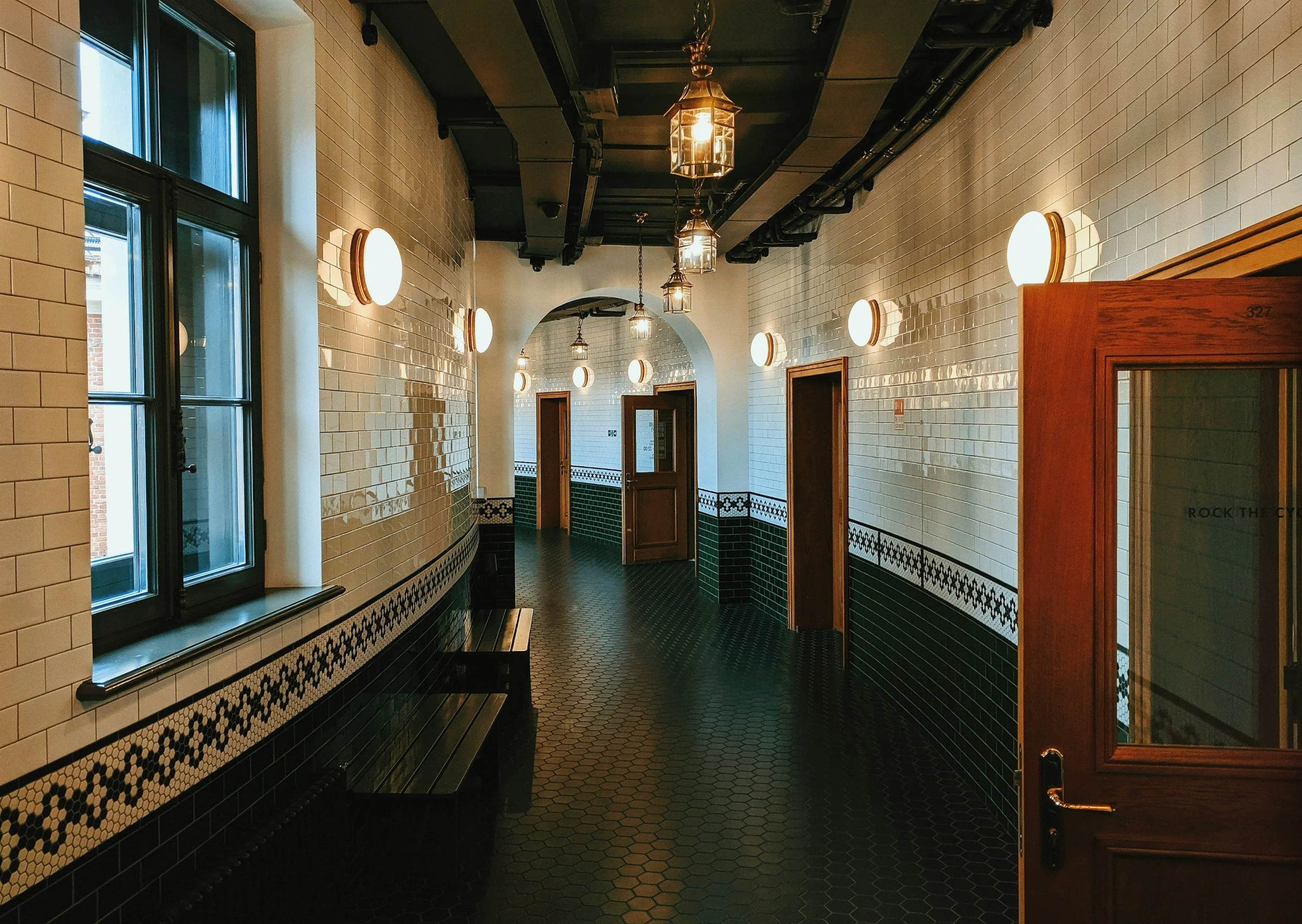 A hallway in a restaurant or café featuring white subway tile walls, black hexagon floor tiles, wooden door frames, circular wall-mounted lights, and vintage-style hanging lanterns illuminating the ceiling.