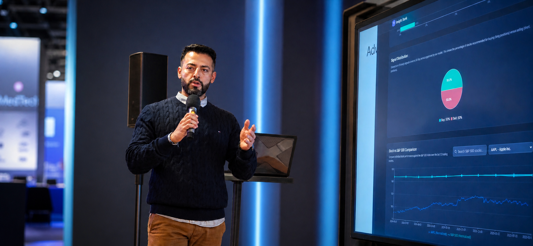 A man with a beard and dark hair giving a presentation in front of large monitors displaying financial graphs and data, holding a microphone and wearing a dark sweater.