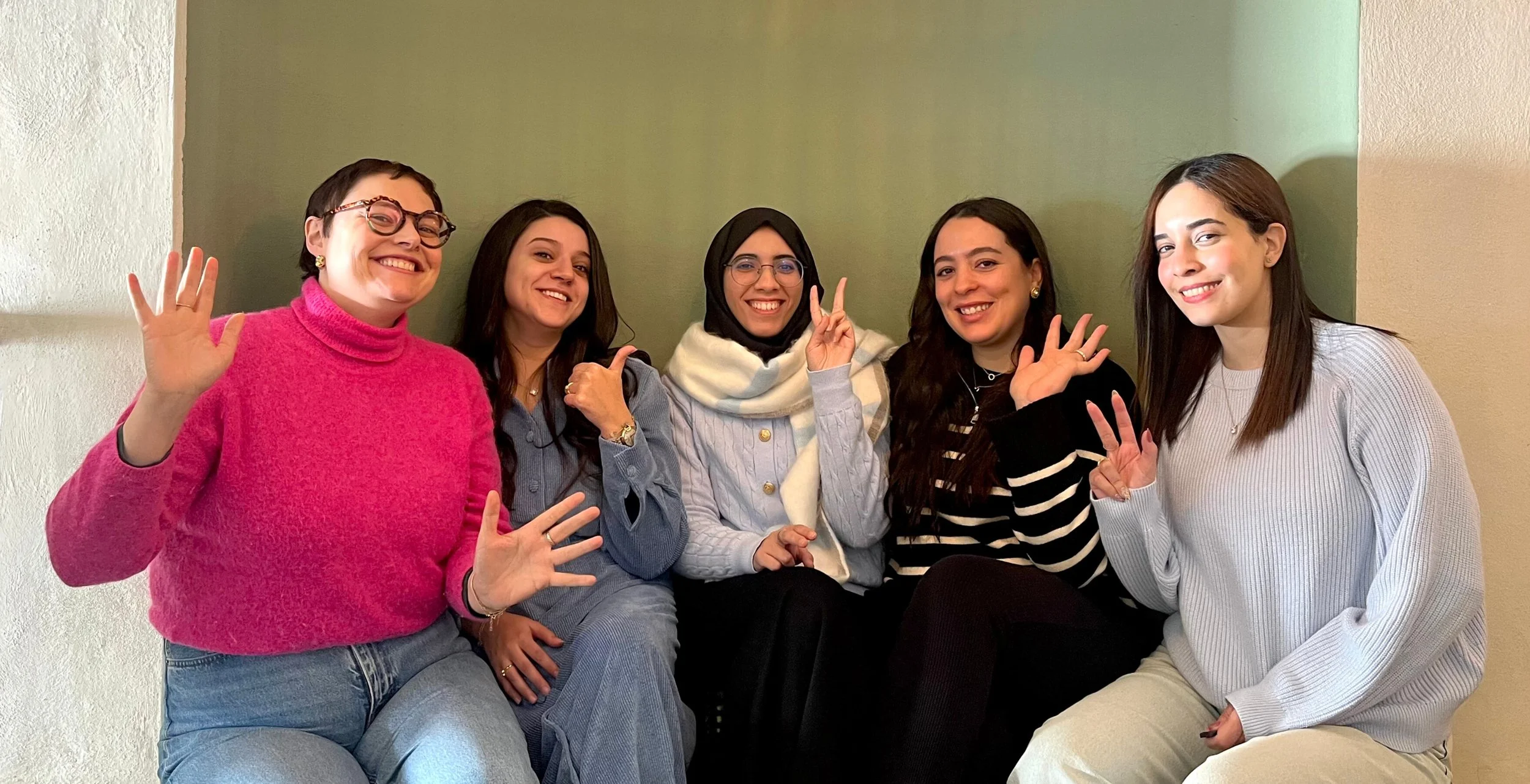 Six women sitting on a bench smiling and waving at the camera, against a two-tone wall.
