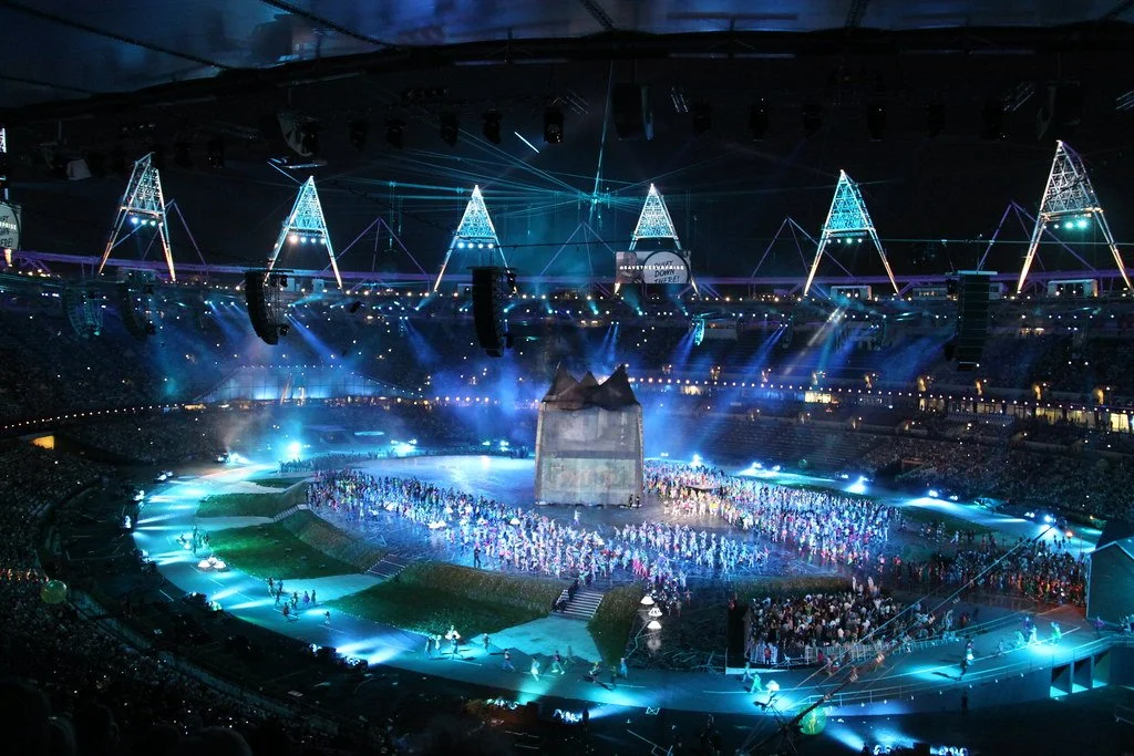 A large stadium with a vibrant performance taking place at night. The scene is illuminated with colorful lights, and a crowd surrounds a central stage area. Performers are dressed in various costumes, and the stadium is filled with spectators.