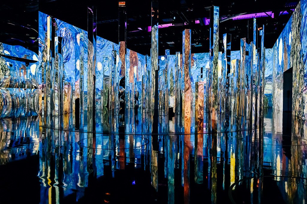 Immersive art installation featuring tall reflective pillars with blue and multicolored light projections, creating a surreal environment with distorted images and reflections on the floor.