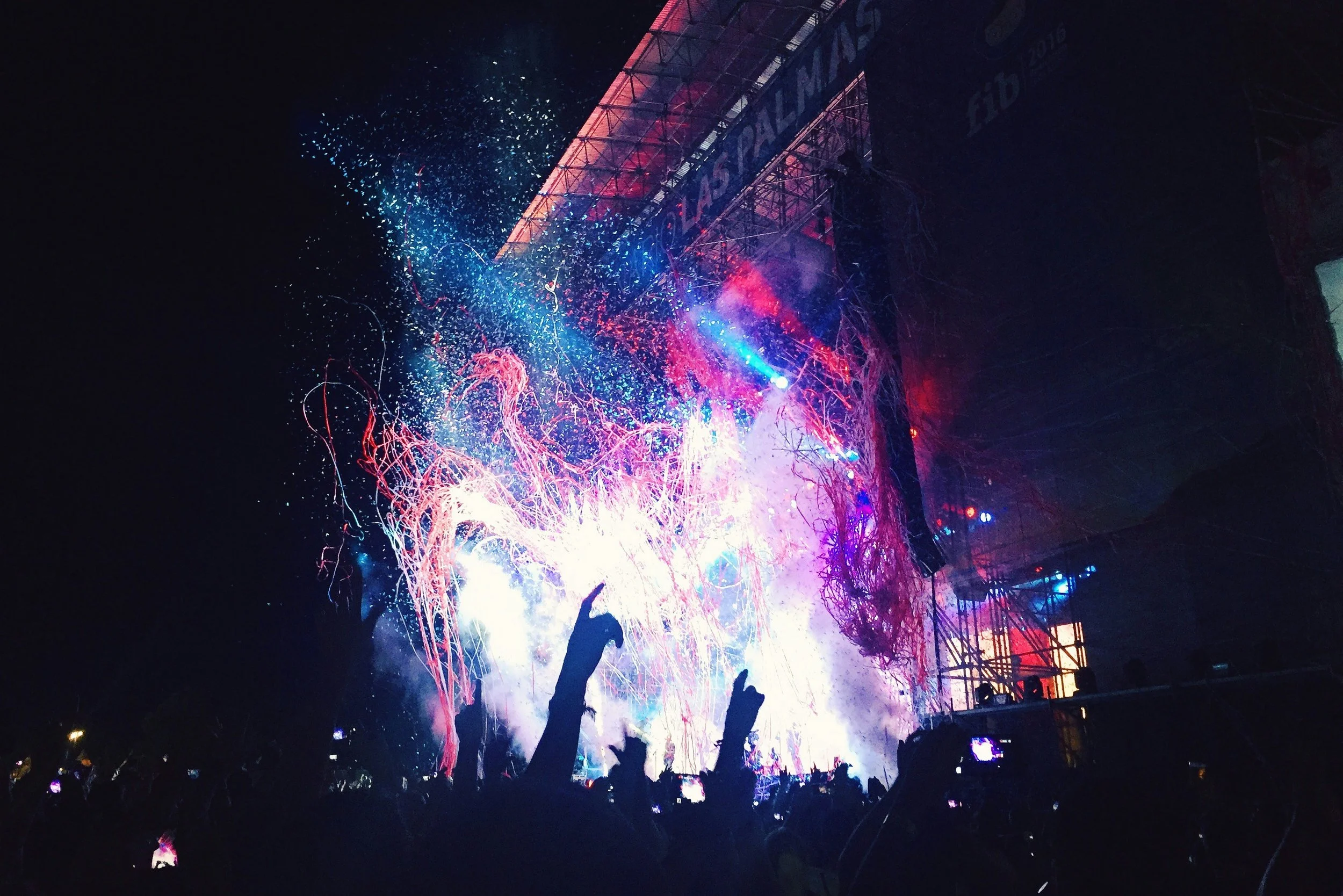 Concert stage with colorful lights and confetti, crowd with raised hands.