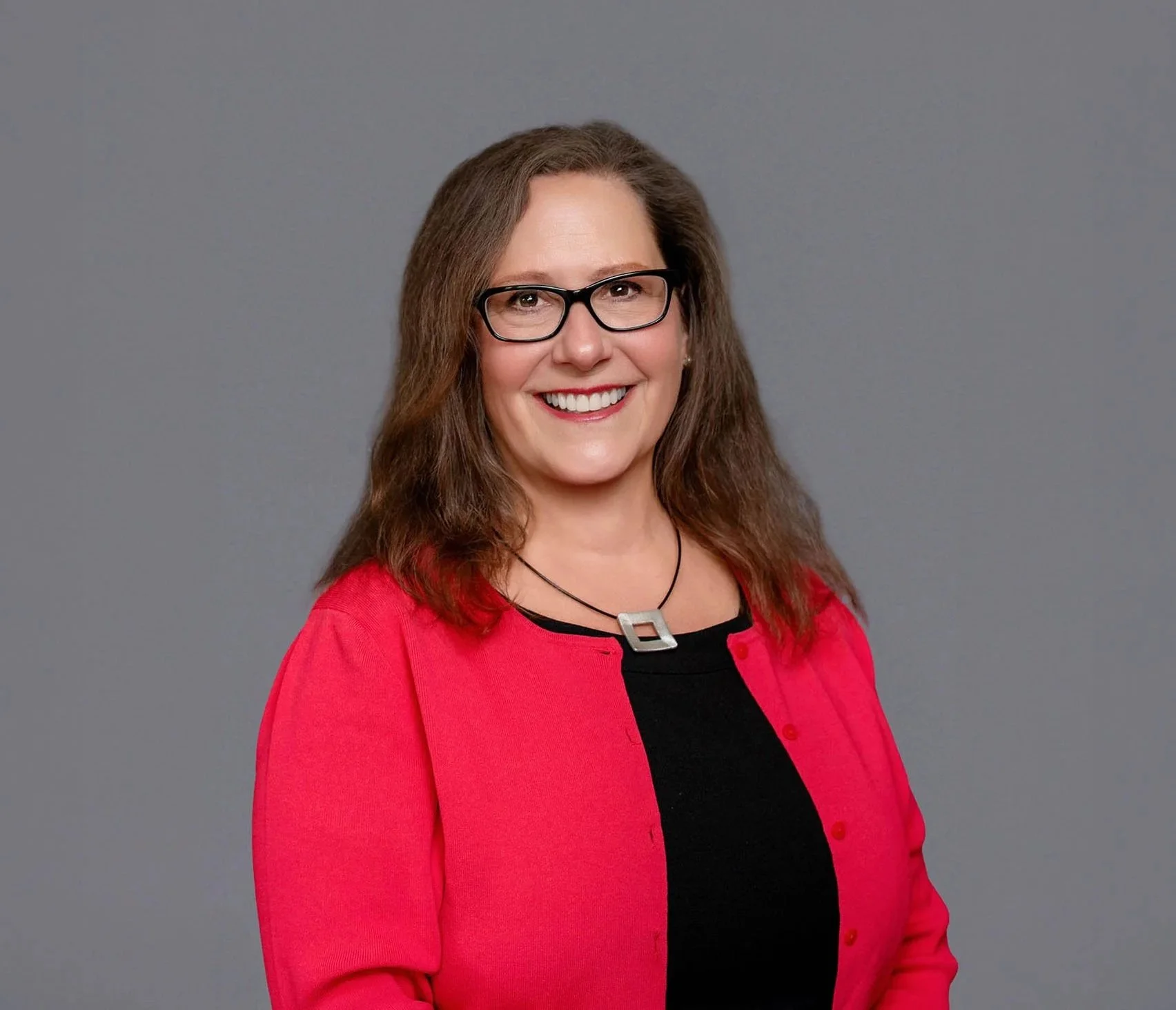 Trisha Mayhew, attorney for family law in Portland Oregon with long brown hair, glasses, and a bright smile wearing a red blazer over a black top, standing against a gray background.