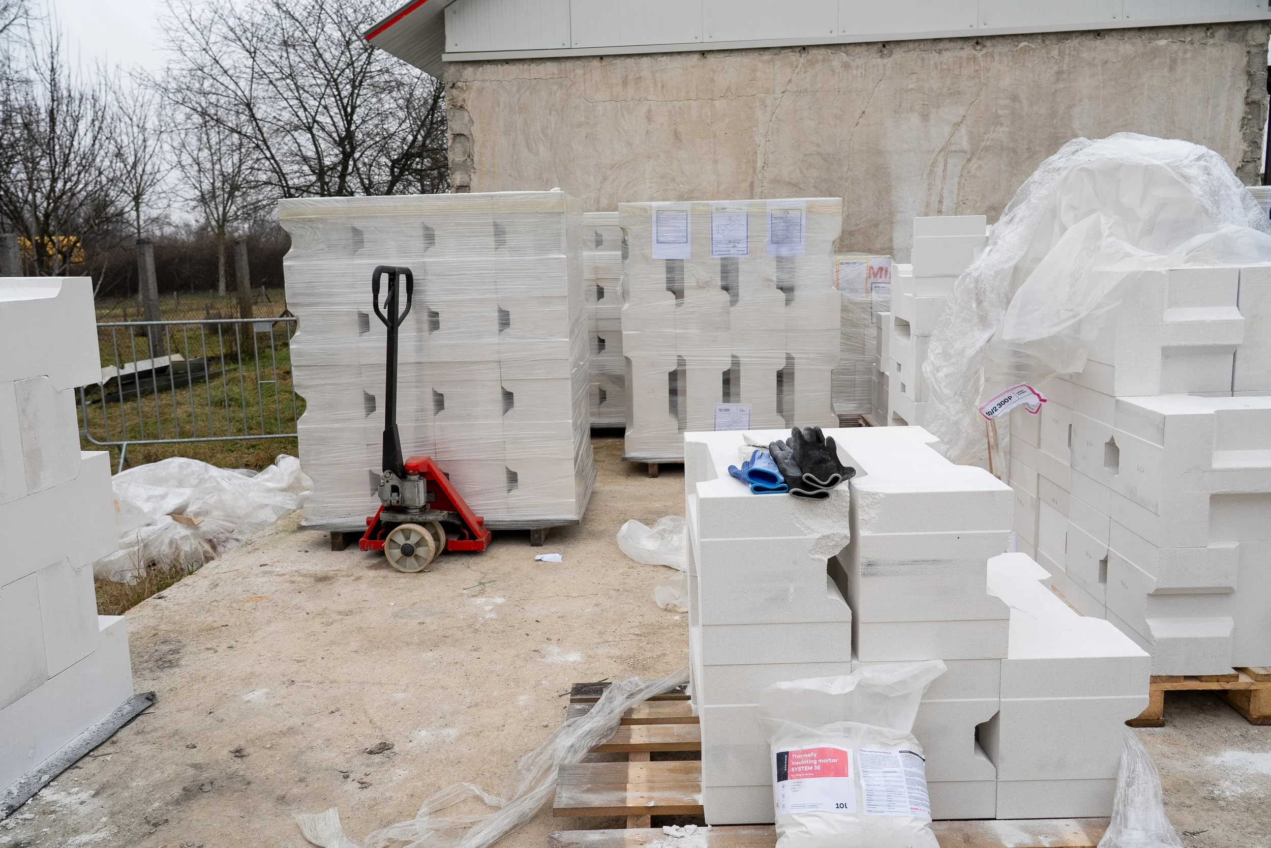 Construction site with white styrofoam blocks, pallets, plastic wrap, gloves, and tools, set against an unfinished building wall and leafless trees in the background.