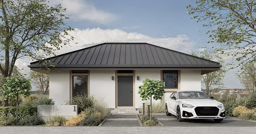 A modern single-story house with a black metal roof, white walls, and wooden window frames. A white Audi car is parked in the driveway, and there are trees and shrubs around the house. The sky is partly cloudy.
