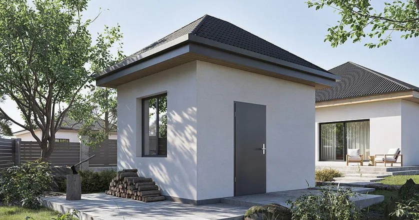 A small, modern white shed with a gray door and black window, surrounded by a garden with trees and grass, in front of a larger house with outdoor seating.