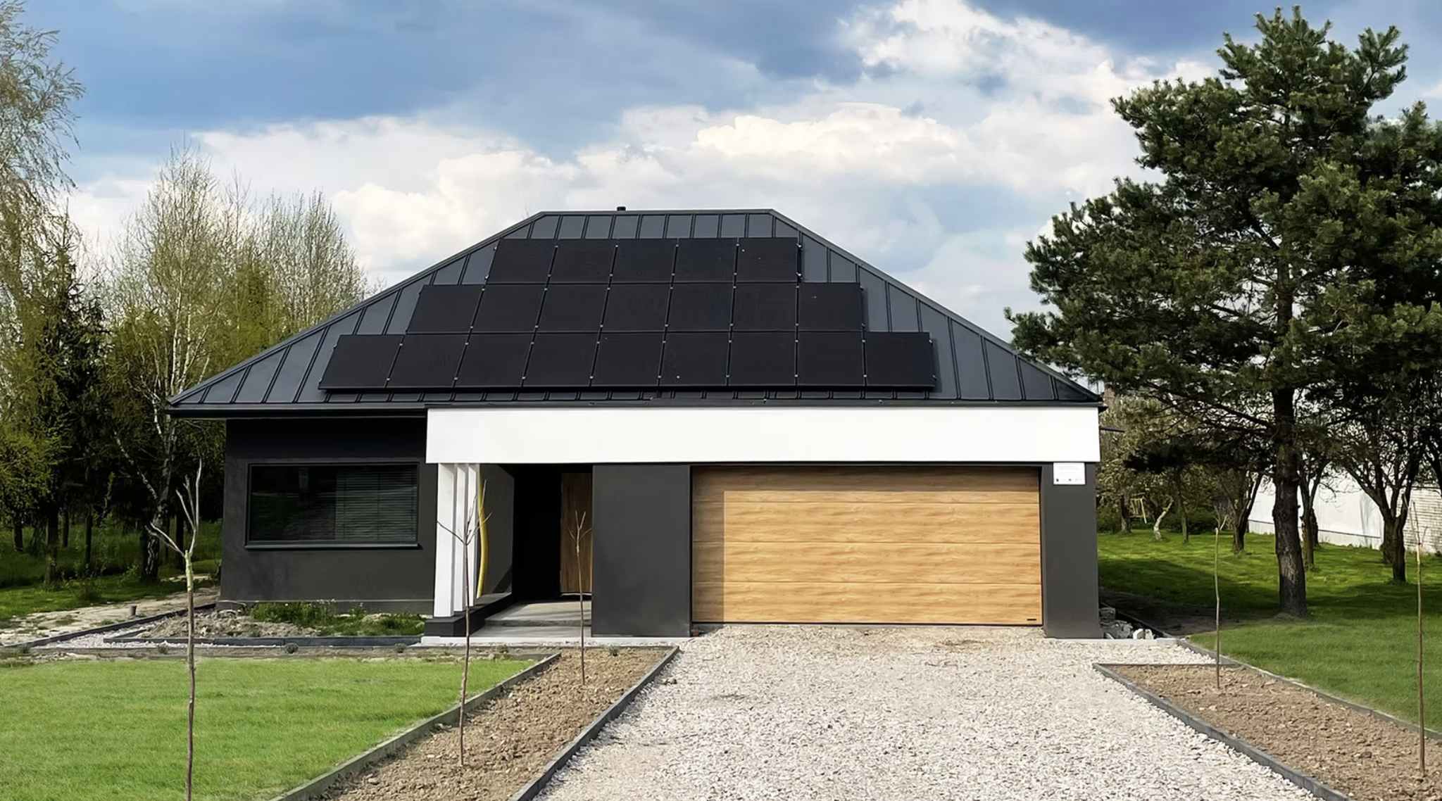 Modern house with black roof solar panels, wooden garage door, surrounded by green trees and grass, cloudy sky.