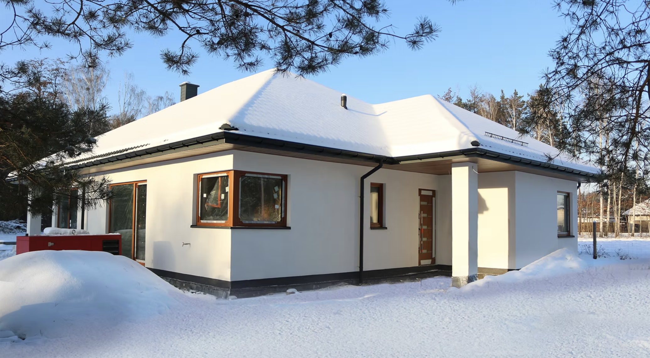 A modern white house with a snow-covered roof, surrounded by snow and trees, under a clear blue sky.