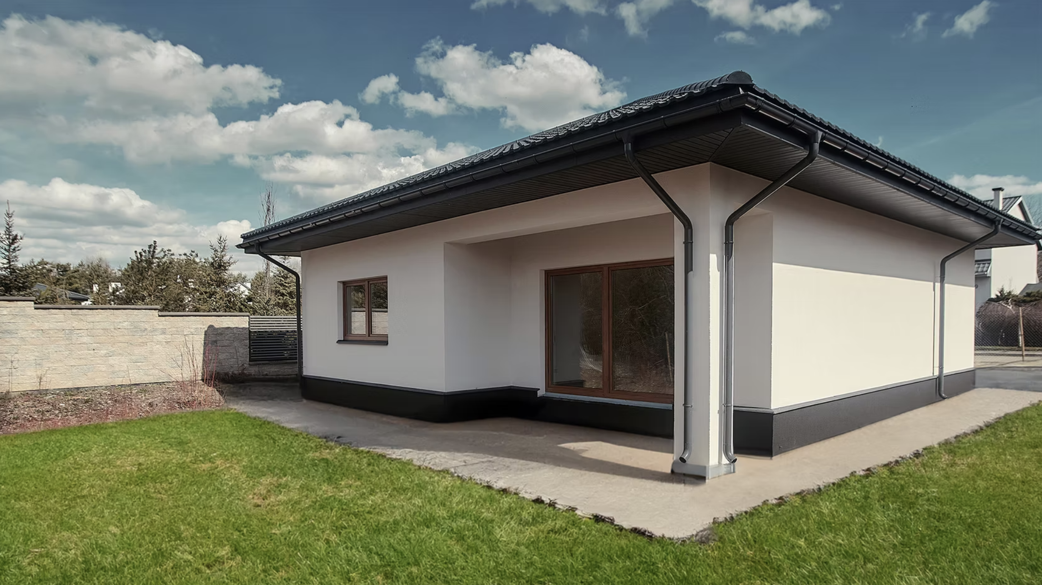 A modern house with white walls, large window and sliding glass door, black roof with gutters, situated on a grassy lawn under a partly cloudy sky.