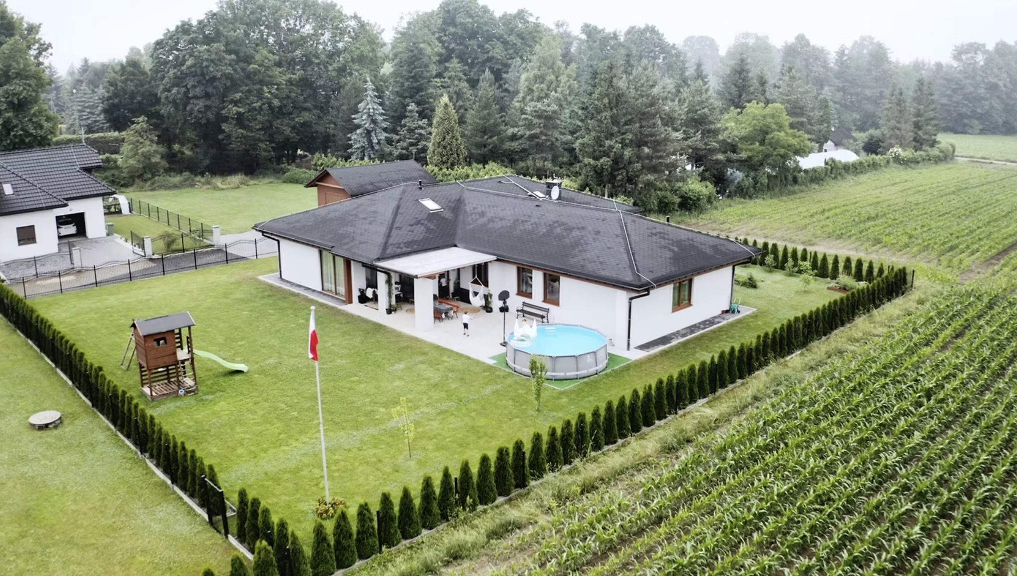 A house with a black roof and a patio, surrounded by a well-maintained lawn, a fenced yard, a children's play area, and a small pool. Trees and fields are visible in the background.