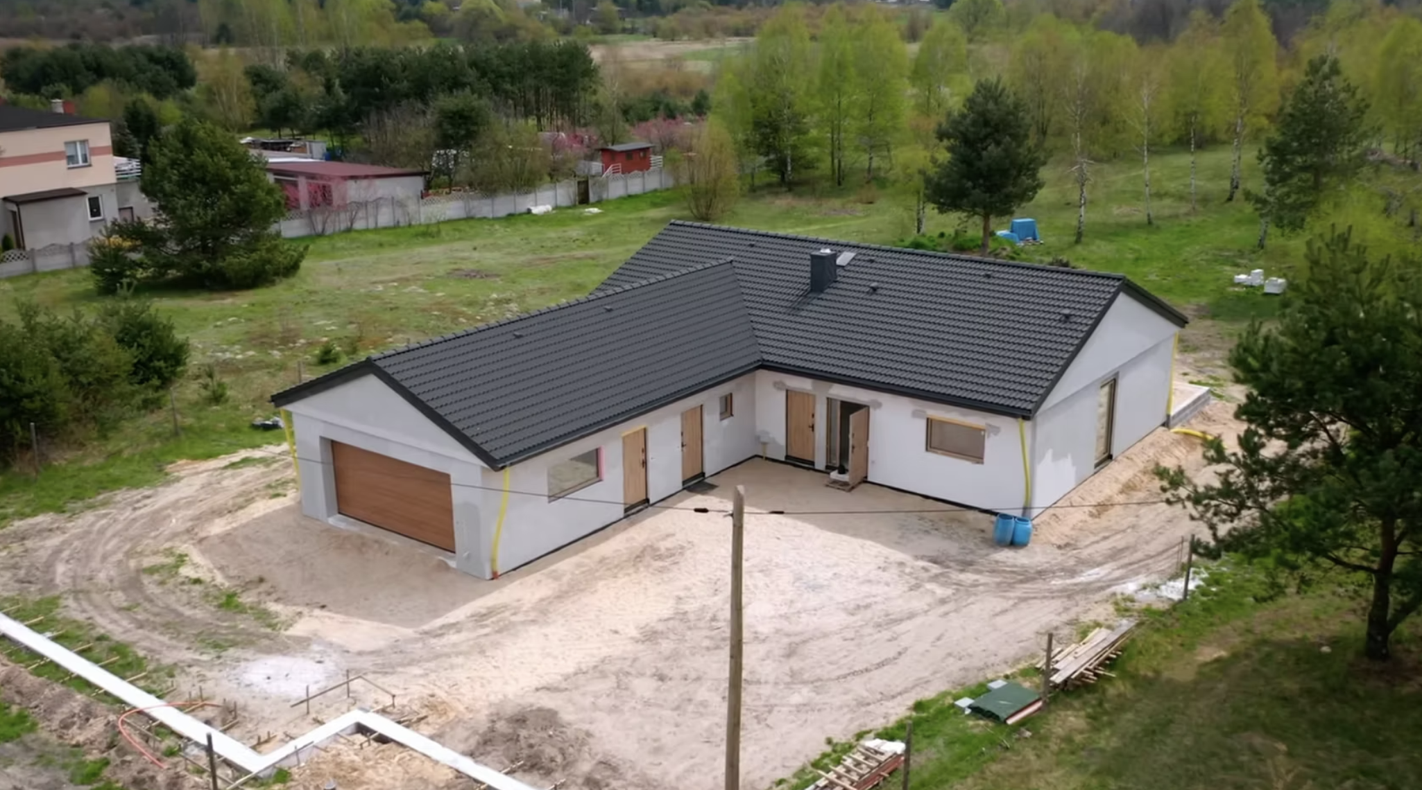 A house under construction with a dark gray tiled roof and white exterior walls, surrounded by green grass and trees, with construction materials and equipment nearby.