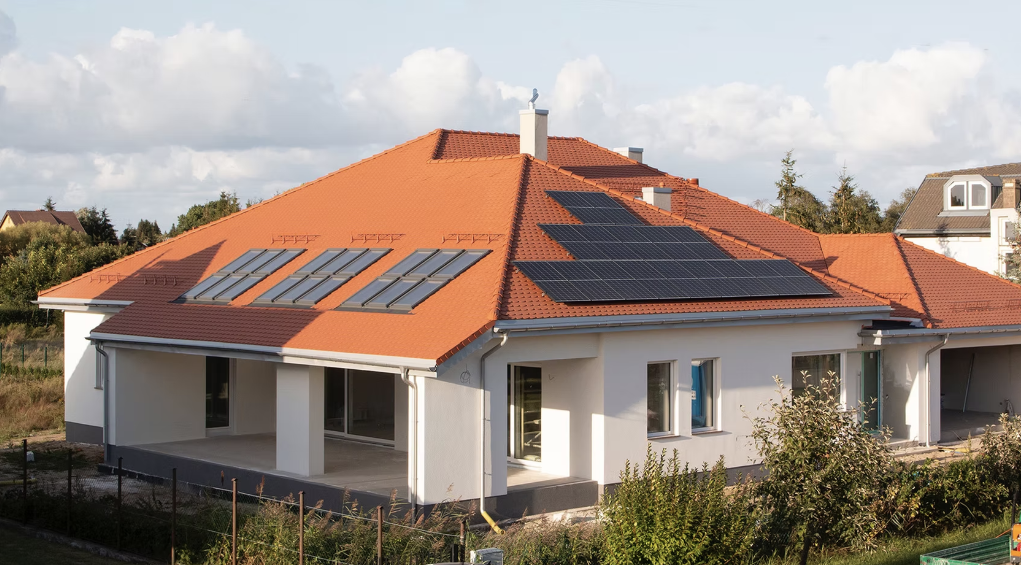 A house with a red-tile roof fitted with multiple solar panels, set against a partly cloudy sky and neighboring houses.