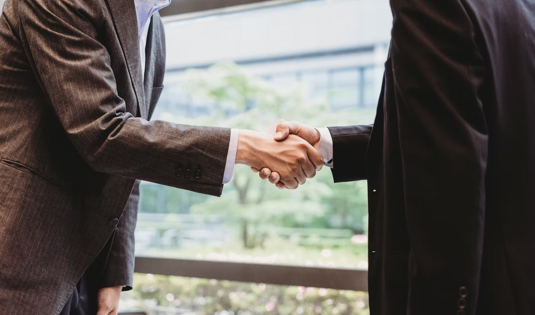 Two men in business suits shaking hands indoors with a window view of trees and buildings in the background.