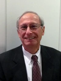 A smiling middle-aged man in a suit and tie, wearing glasses, standing against a plain wall.