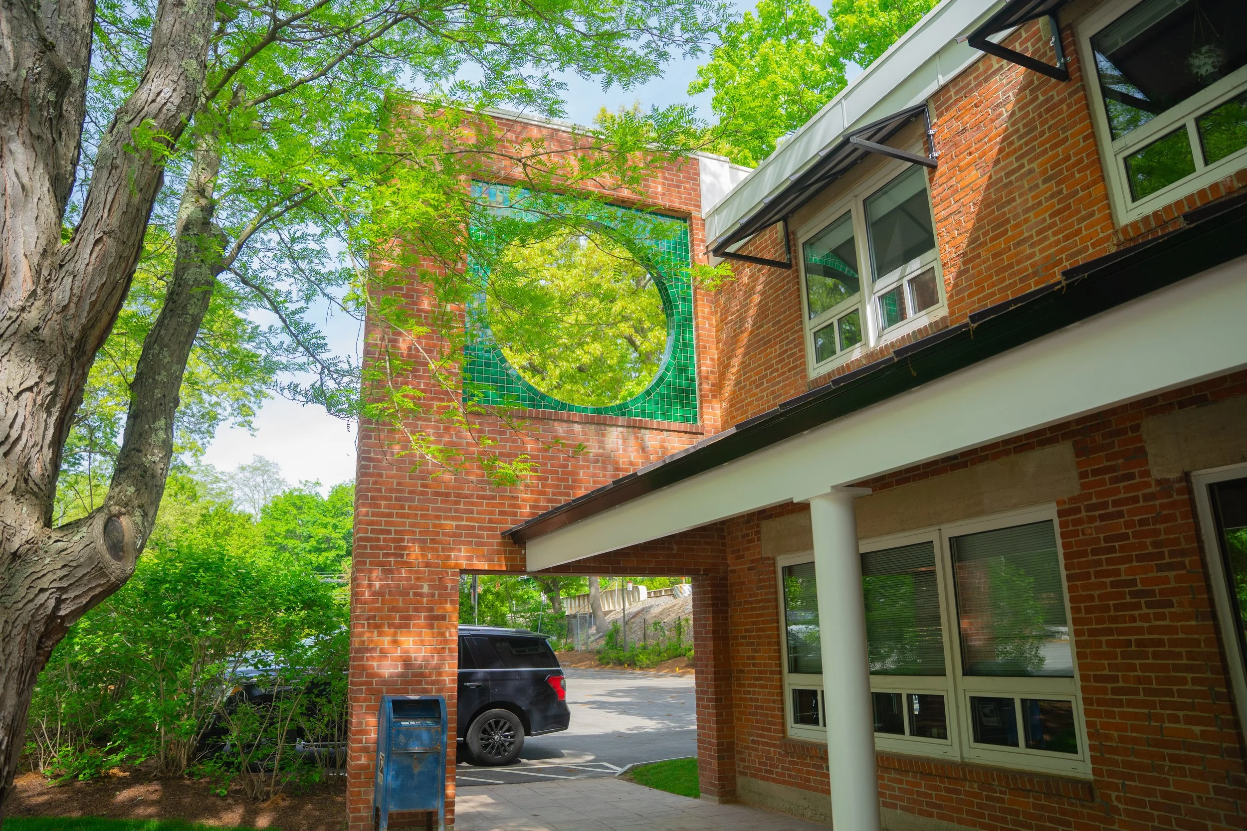 Backyard view showing a brick house with large windows, a car parked in the driveway, and a lush green tree with bright leaves. A mosaic art piece is visible on the house wall. There is a trash bin on the sidewalk.