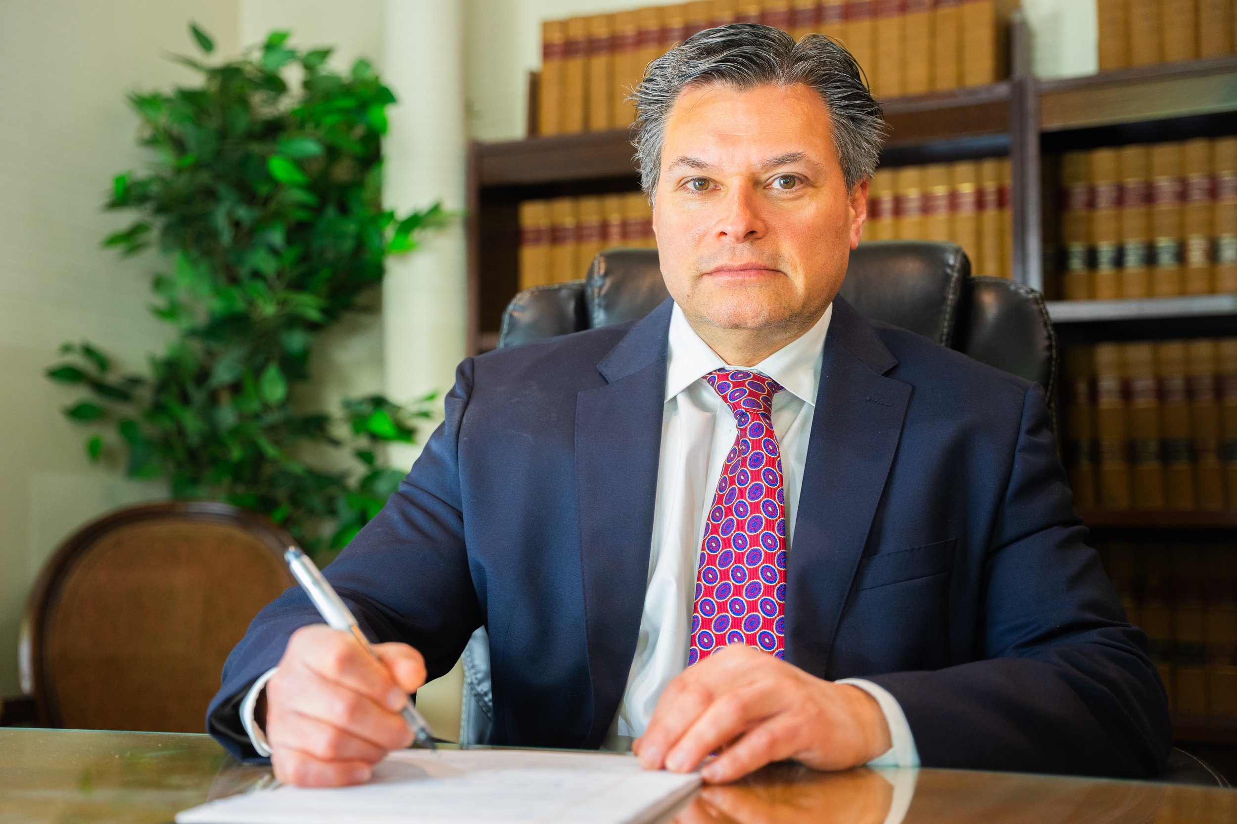 A man in a blue suit and colorful tie writing on paper at a desk in an office with bookshelves and a plant in the background.