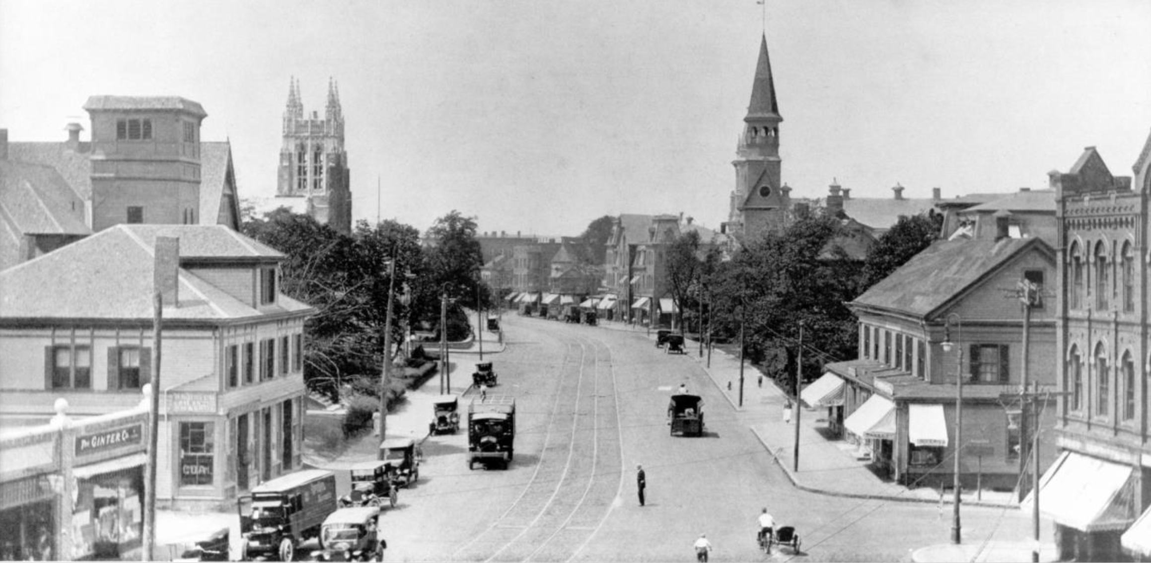 Historical black-and-white photo of early 1900's downtown Newton, Massachusetts with early automobiles, trees, multi-story buildings, and church steeples in the background.
