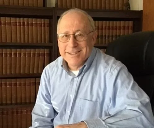 An older man with glasses, smiling, wearing a light blue shirt, sitting at a desk in front of a bookshelf filled with books.