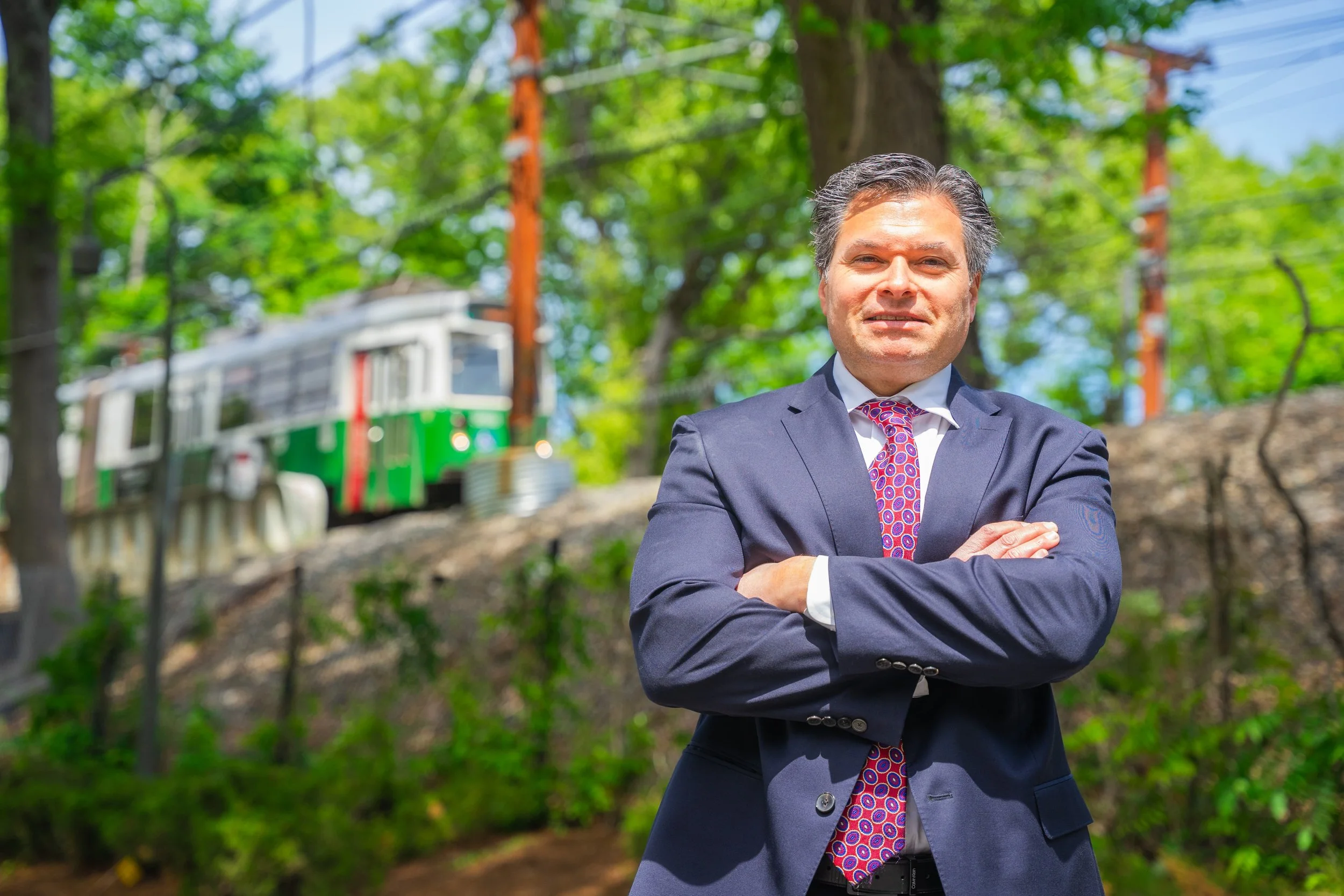 A man wearing a suit with a patterned tie, standing outdoors with arms crossed, in front of trees and a blurred green and white train in the background.