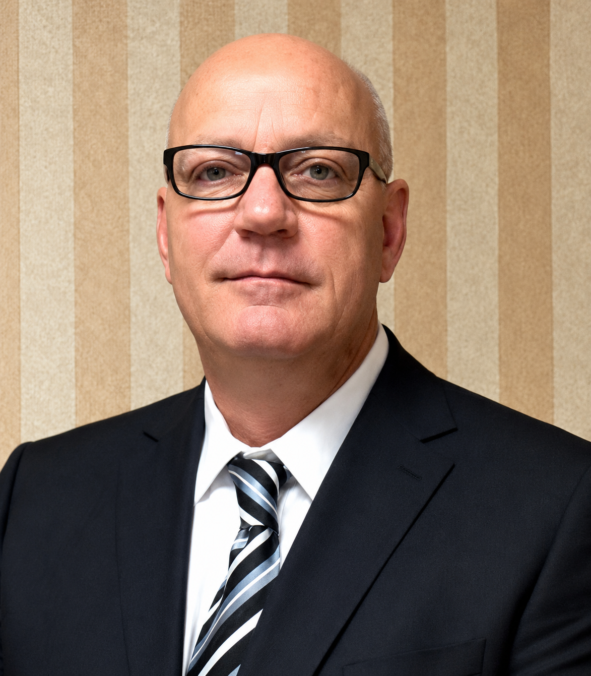 A bald man wearing glasses, a dark suit, white shirt, and striped tie, looking directly at the camera against a beige-striped wallpaper background.