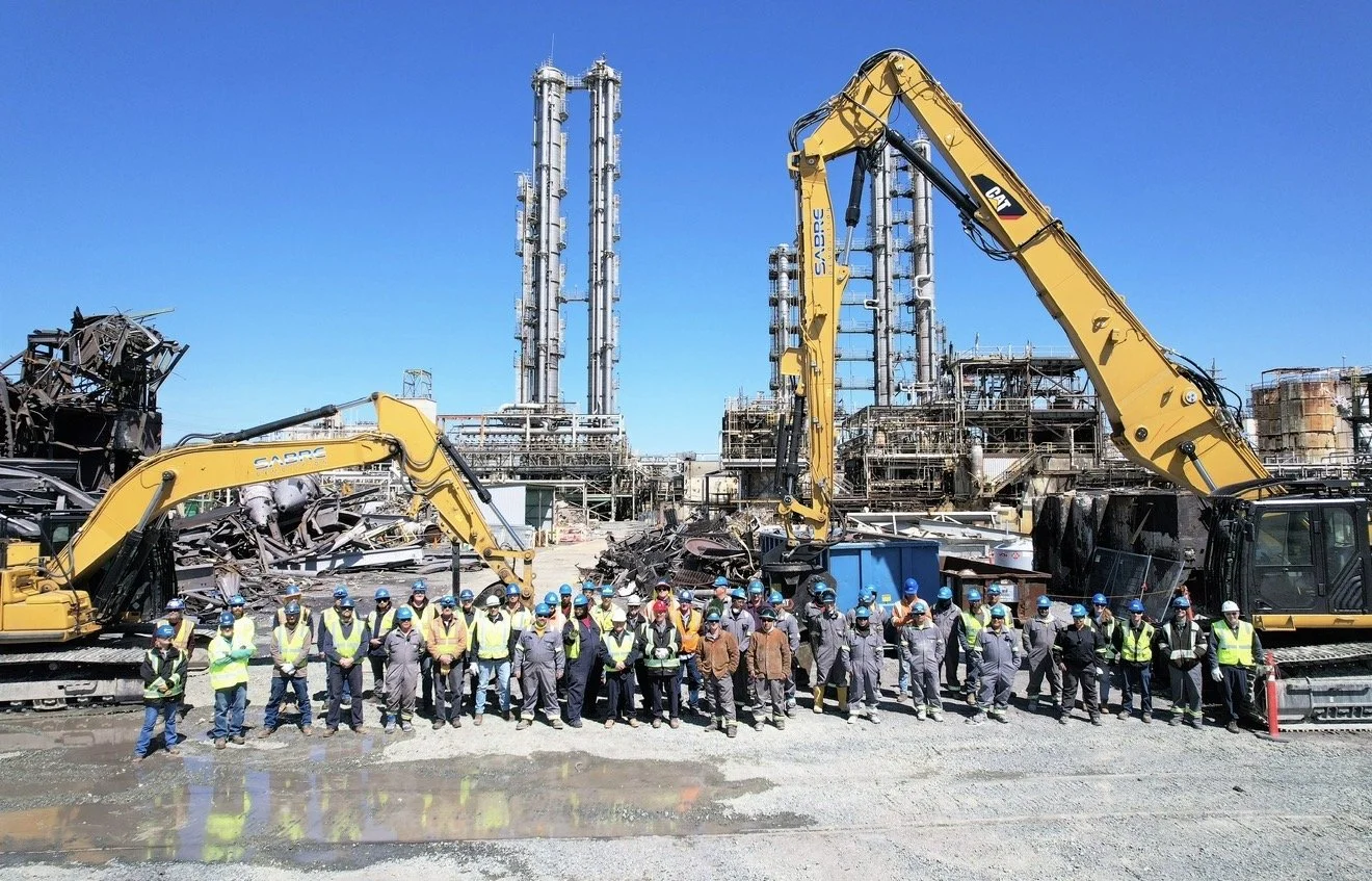 Group of construction workers wearing safety helmets and vests standing in front of construction equipment at an industrial site with factories or refineries in the background under a clear blue sky.