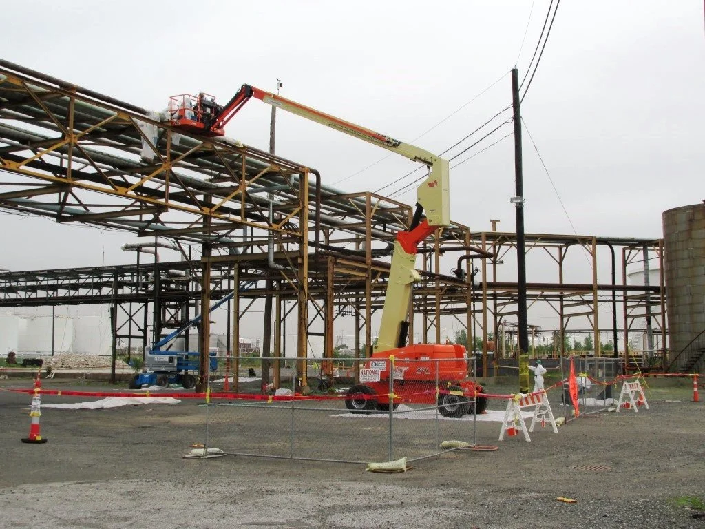 Construction site with a crane lifting workers on a scaffolded industrial structure, fenced off with safety barriers and cones.