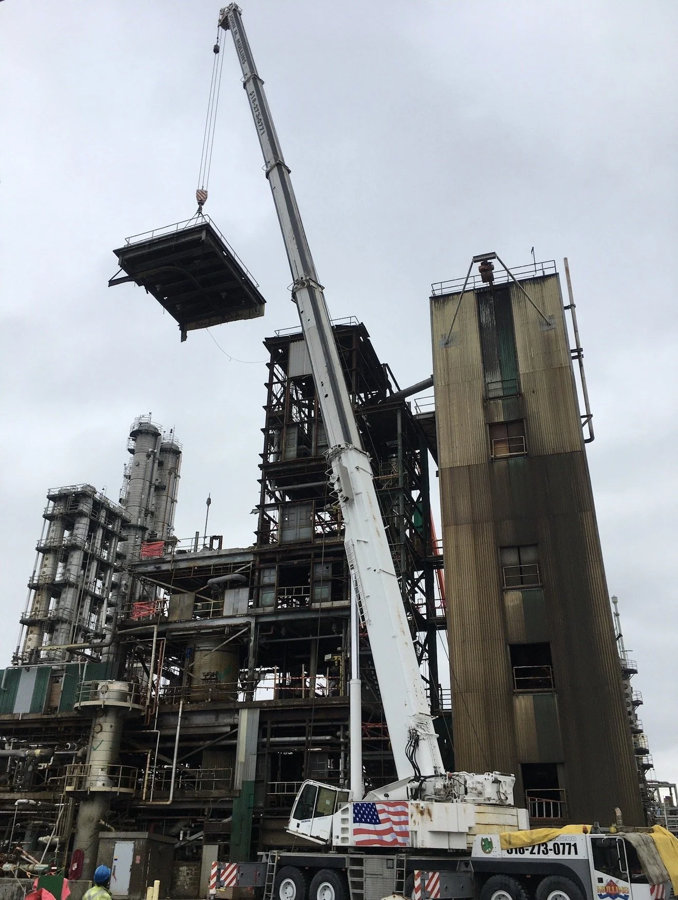 A large construction crane lifting a platform at an industrial site with factory buildings and pipes in the background.