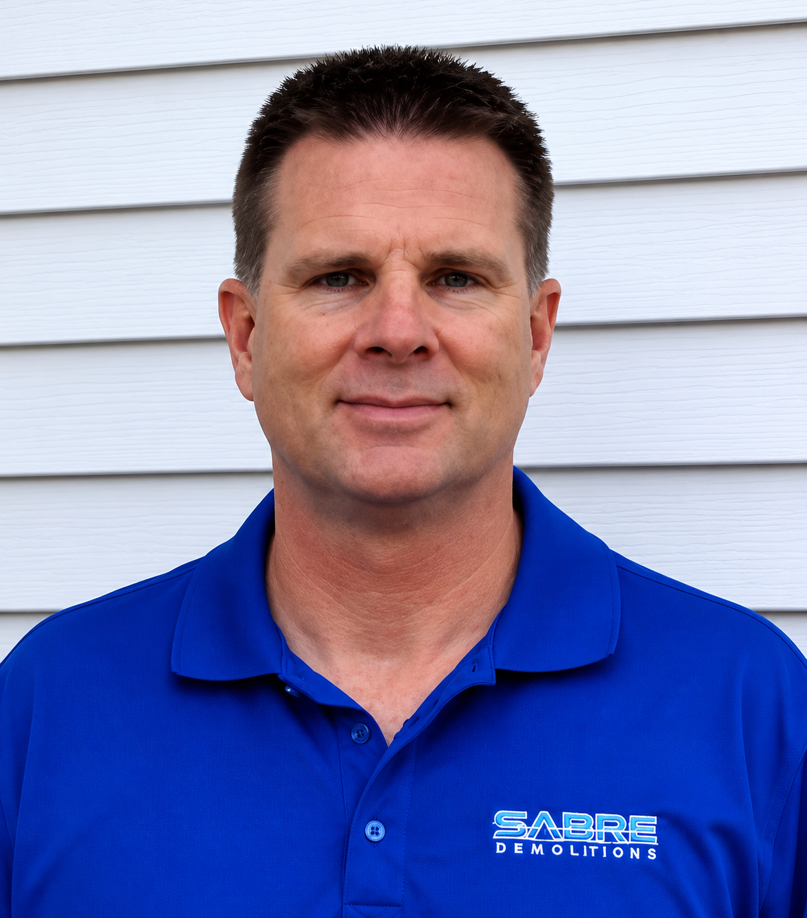 A man with short dark hair wearing a blue polo shirt with "SABRE DEMOLITIONS" logo, standing in front of white horizontal siding.