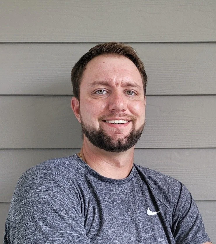 A smiling man with short brown hair and a beard, wearing a gray Nike athletic shirt, standing against gray wooden siding.