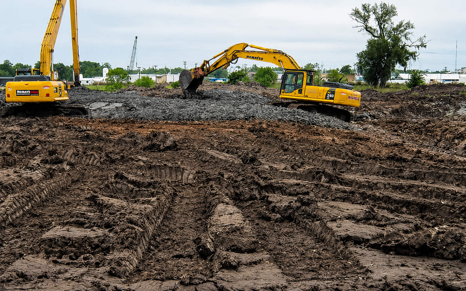 Construction site with yellow excavators moving dirt and gravel, with tire tracks in the foreground and trees and buildings in the background.