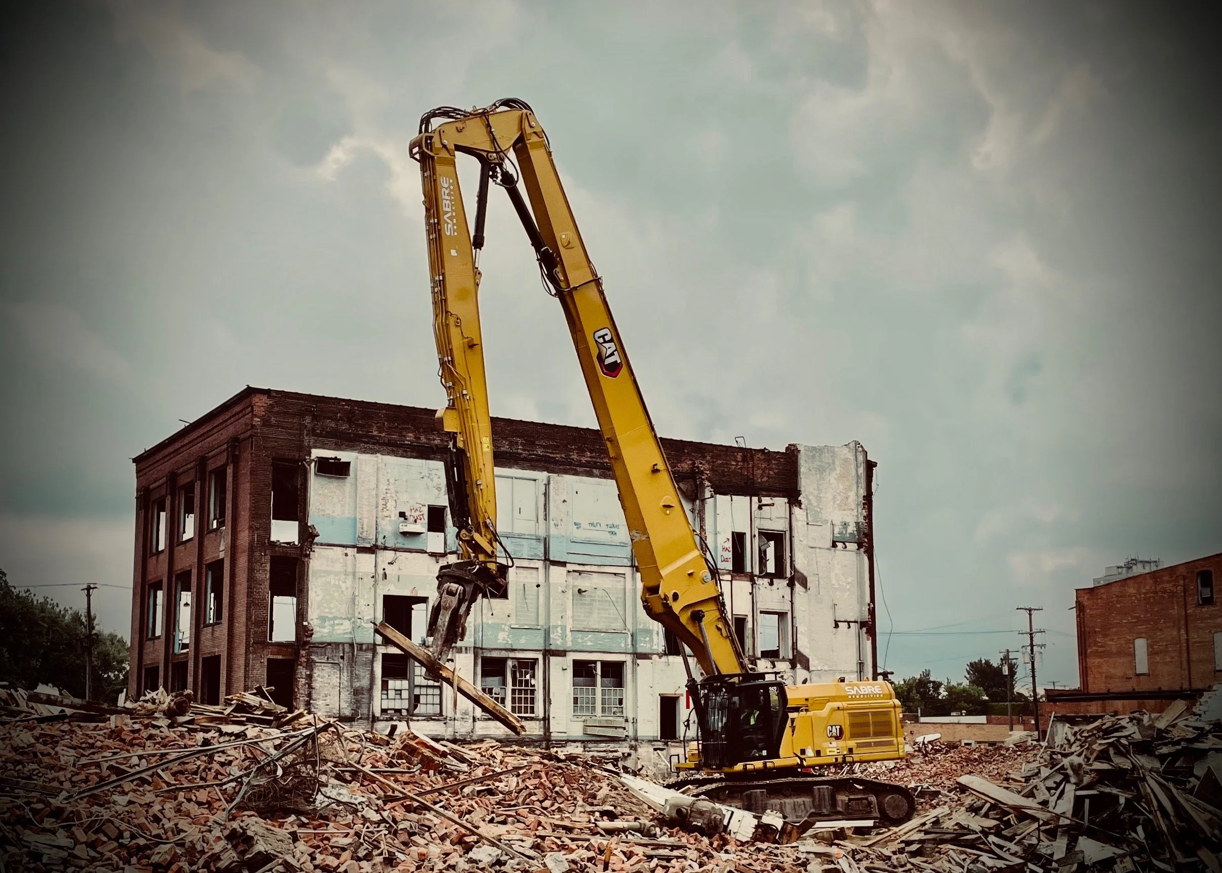 Construction excavator tearing down a brick building, piles of debris in the foreground, cloudy sky.