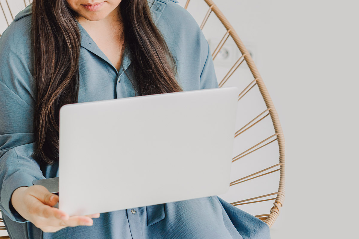 Woman with long dark hair in blue shirt sitting in modern rattan chair, working on a white laptop.