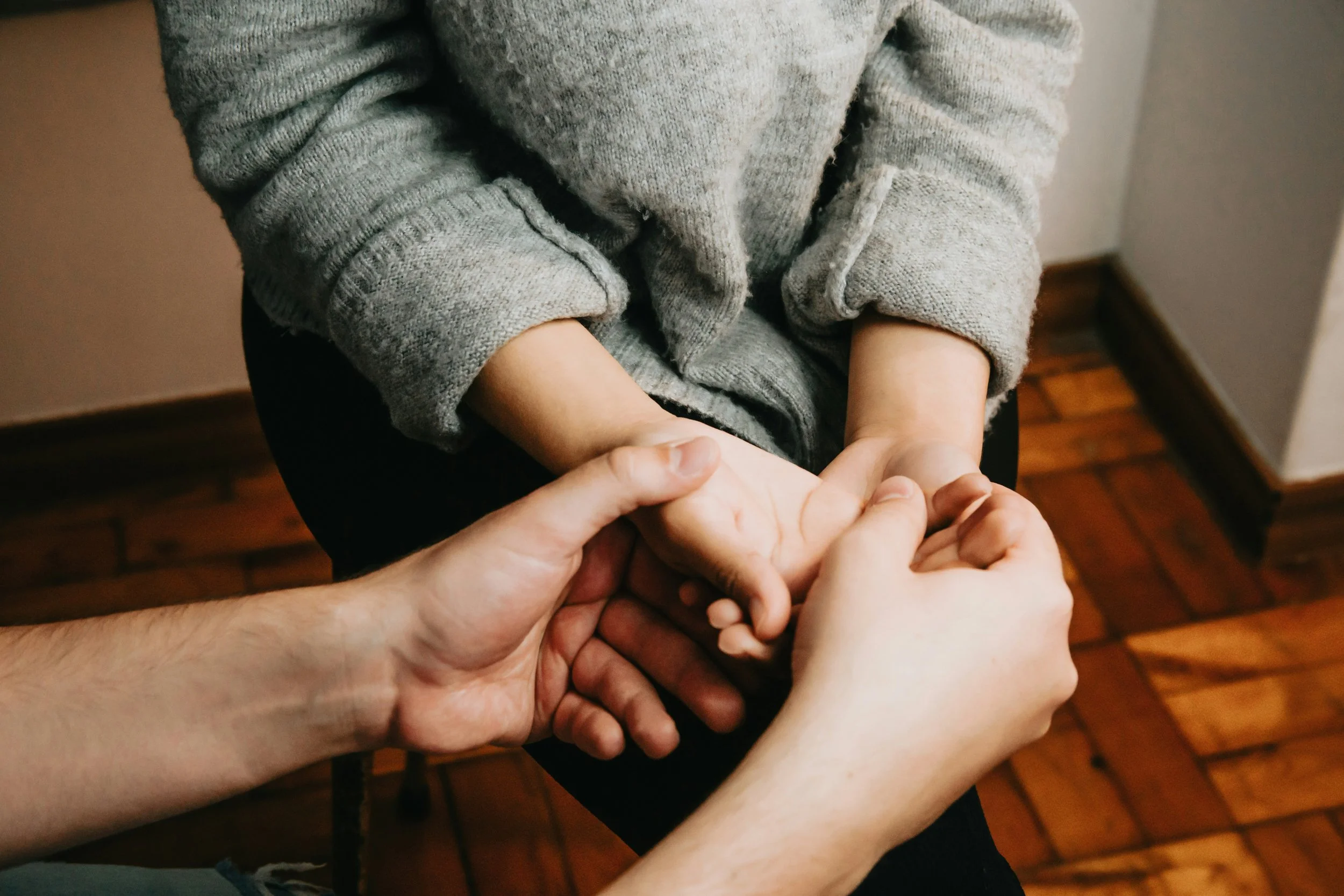 Multiple hands holding a small child's hand indoors with wooden flooring and a beige wall.