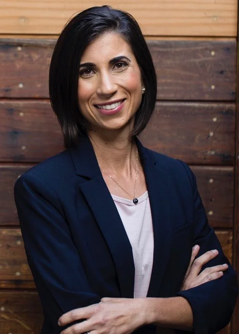 A woman with shoulder-length dark hair and a friendly smile, wearing a navy blazer over a light blouse, standing in front of a wooden wall.