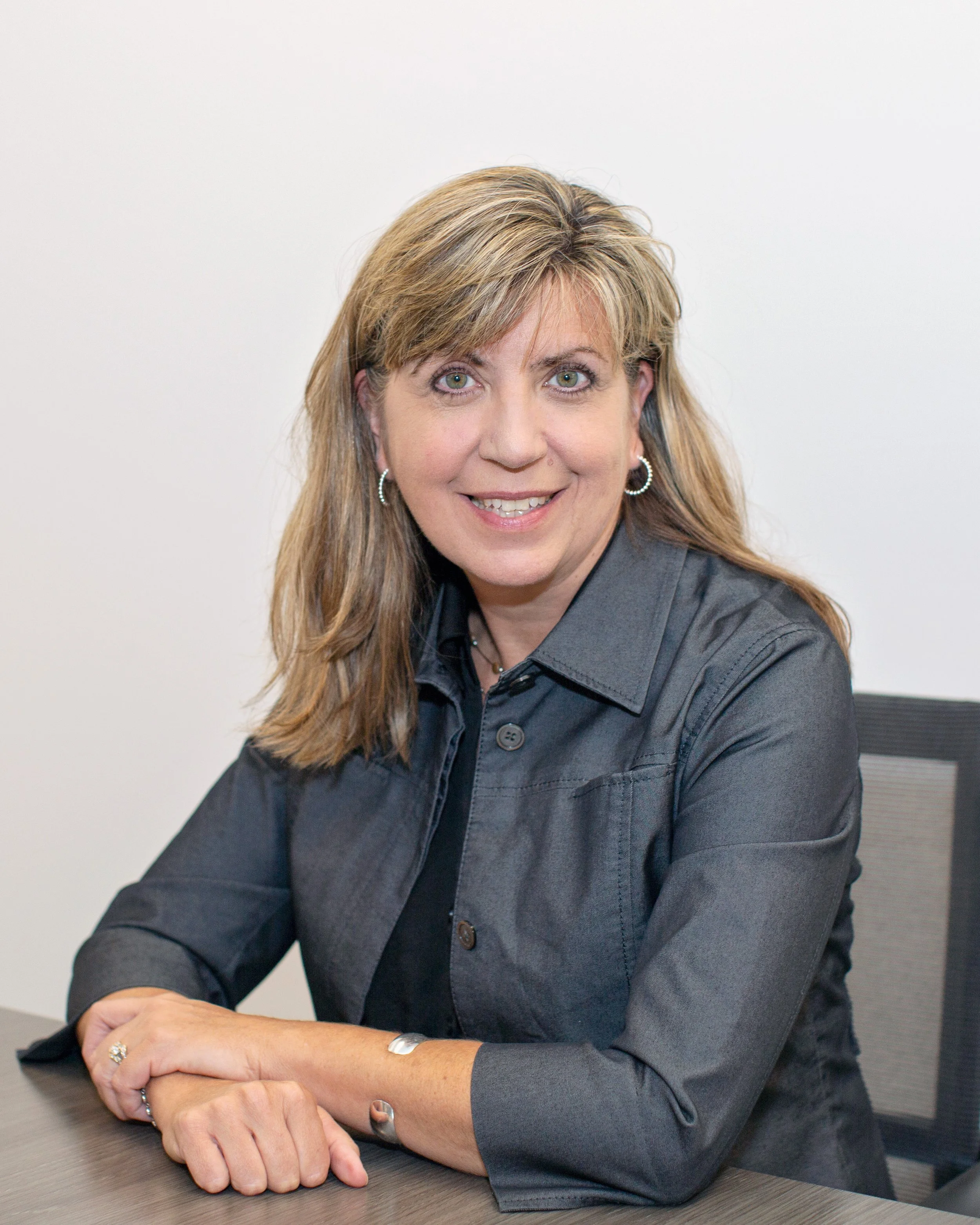 A woman with shoulder-length, light brown hair, wearing a dark gray blazer and small hoop earrings, sitting at a table and smiling at the camera.